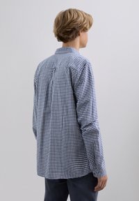 Persona con cabello rubio corto que lleva una camisa de cuadros azul y blanca de manga larga, de pie de espaldas contra un fondo sencillo.