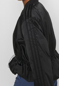 Svart bomberjacka i glänsande nylon, med elastisk midja, ribbad krage och tre svarta ränder längs ärmarna.