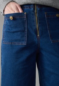 Denim pants with a deep blue color, featuring two front pockets, brass buttons, and a gold zipper. Contrast stitching adds texture.