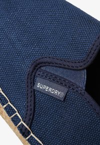 Zapatilla de lona azul marino con parte superior texturizada, borde cosido y una etiqueta de Superdry visible cerca de la costura. Suela hecha de material de cuerda natural.