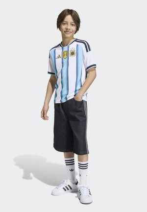 Chłopiec stoi z ręką w kieszeni, ubrany w koszulkę argentyńskiej drużyny piłkarskiej, czarne szorty, białe skarpetki Adidas i białe trampki na jednolitym tle.