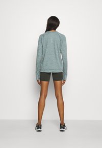Langärmliges, heather-blaues Shirt mit Raglanärmeln, kombiniert mit dunkelgrünen Shorts. Das Modell steht mit dem Rücken zur Kamera und trägt schwarze Sportschuhe.