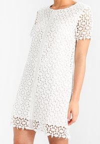 Vit spetsklänning med ett blommigt utskärningsmönster, korta ärmar, rund halsringning och vågig nederkant med intrikata detaljer.