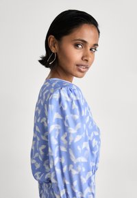 Bluză de culoare bleu deschis cu un model abstract alb, având mâneci bufante, guler rotund și un material moale, texturat. Design elegant și simplu.