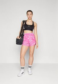 Juoda sportinė liemenėlė su trimis baltomis juostelėmis, rožinės rožių dažų šortai, balti sportiniai bateliai ir juoda pečių krepšys. Matosi dryžuotos pėdkelnės.