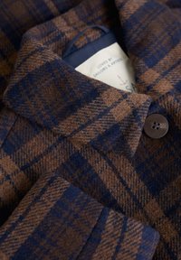 Zbliżenie na kolorowy brązowo-granatowy płaszcz w kratę, z kołnierzem, ciemnym guzikiem i częściowo widoczną białą metką w środku.