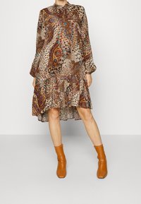 Le mannequin porte une robe longue à motifs avec un ourlet asymétrique et des manches bouffantes, associée à des bottines marron, sur un fond uni.