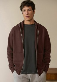 Burgunderfarbene Zip-Hoodie mit Fronttasche und Kapuze, getragen über einem dunkelgrauen, eng anliegenden Hemd. Glattes Material mit lässiger Passform.