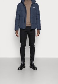 Veste matelassée bleu marine avec fermeture éclair et capuche, portée sur un pull marron. Jean noir slim et bottines noires brillantes complètent le look.