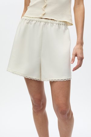 Kvinne iført høytlivede kremfargede satengshorts med elastisk midjebånd og blonderkant nederst, kombinert med en kremfarget ermeløs knappet topp.