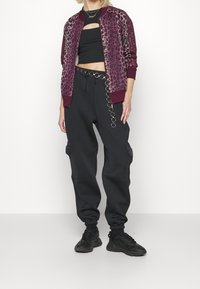 Bordová bomber bunda s leopardím vzorem a černý crop top, doplněné vysokým pasem černých tepláků s řetízkovým detailem a bočními kapsami.