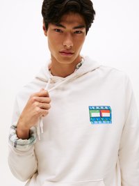 Hanorac crem deschis, cu un patch colorat pe piept, însoțit de manșetele interioare cu model în carouri și șnururi de ajustare. Material texturat.