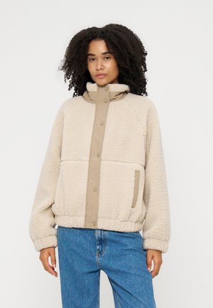 Femme aux cheveux bouclés portant une veste polaire beige avec des boutons sur le devant et un jean bleu, debout devant un fond uni.