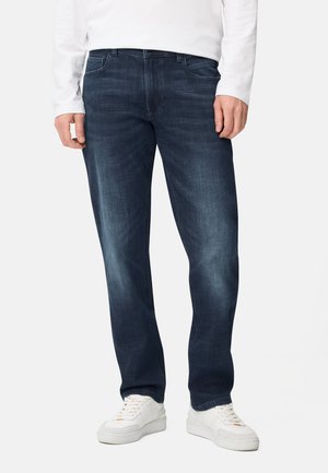 Mand iført mørkeblå jeans med subtilt falmet look, hvid langærmet skjorte og hvide sneakers, stående med hænderne afslappet ned langs siderne.