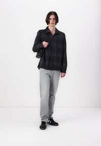 Svart skjorte med knappelukking og rutete mønster, grå jeans, svarte sneakers og en svart skulderveske; modellen står mot en hvit bakgrunn.
