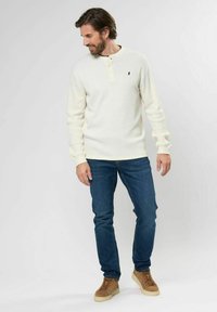 Krémsový žebrovaný pletený dlouhý rukáv henley triko s knoflíkovou légou na tři knoflíky, v kombinaci s modrými džíny a světlým hnědým obuví.