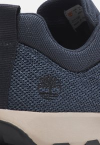 Dunkelblaue Mesh-Sneaker mit schwarzen Akzenten, die mit einer strukturierten Überlagerung und einem Logo, das einen Baum darstellt, ausgestattet sind. Leichtes Design und flexible Sohle.