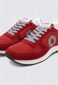 Zapatillas deportivas rojas con acabado suave de ante, cordones grises y suela de goma negra. Presentan un logotipo circular gris en el lateral.