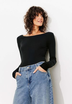 Femme aux cheveux bruns bouclés portant un haut noir à manches longues et un jean bleu ample taille haute, debout contre un mur blanc les mains dans les poches.