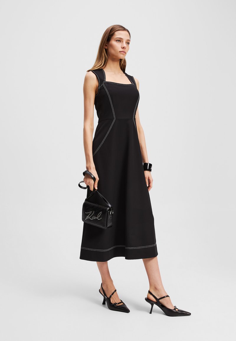 Schwarzes ärmelloses Kleid mit quadratischem Ausschnitt, weißer Naht und A-Linien-Form. Kombiniert mit einer schwarzen Handtasche und spitzen High Heels.