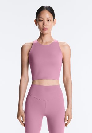 Femeie purtând un top scurt roz pal fără mâneci și colanți cu talie înaltă, stând în fața unui fundal alb simplu.