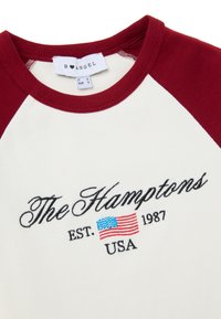 Camisa de manga raglán con cuerpo blanco y mangas burdeos. Presenta el texto bordado "The Hamptons" y una bandera de EE. UU. Material de algodón, cuello acanalado.