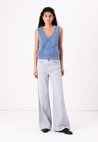 Blauwe gebreide mouwloze top met een V-hals en zigzagpatroon, gecombineerd met lichtgrijze wijde jeans en zwarte schoenen.