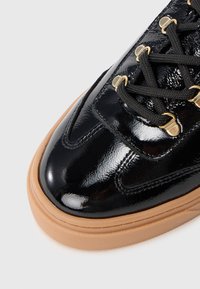 Gros plan sur une sneaker en cuir verni noir avec des lacets noirs, des œillets dorés et une épaisse semelle en caoutchouc beige.