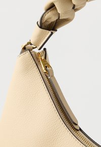 Smėlio spalvos odinė rankinė su faktūruota apdaila, aukso spalvos užtrauktuku ir išskirtine mazguota rankena iš to paties medžiagos.