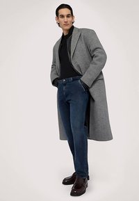 Šedý kabát z vlněné směsi, černý svetr na zip, modré džíny a hnědé kožené boty. Model stojí s rukama v kapsách, což vytváří uvolněný vzhled.