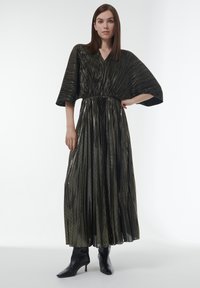 Robe longue plissée noire avec des rayures dorées métalliques, présentant un décolleté en V et des manches larges, accompagnée de bottes à talons noires.