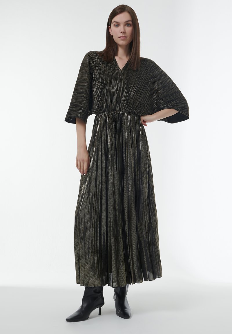 Robe longue plissée noire avec des rayures dorées métalliques, présentant un décolleté en V et des manches larges, accompagnée de bottes à talons noires.