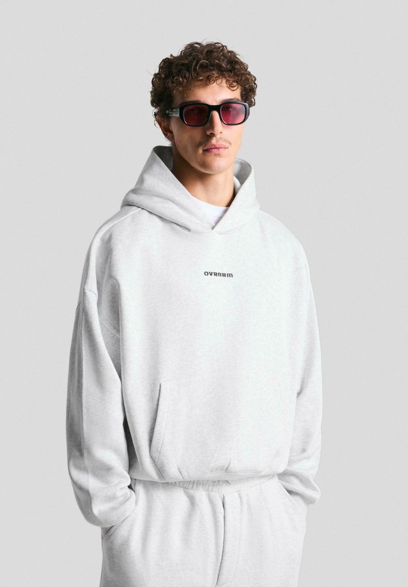 Felpa grigia con tasca frontale, caratterizzata da un piccolo logo nero. Indossata con pantaloni della tuta abbinati e occhiali da sole alla moda. Texture liscia.