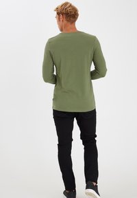 Mand med kort lyst hår iført en olivengrøn langærmet skjorte, sorte bukser og sorte sneakers, står med ryggen til mod en hvid baggrund.