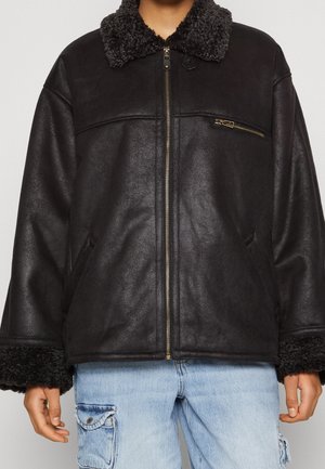 Zwarte shearling jas met ritssluiting en een getextureerde kraag en manchetten, gedragen over lichtblauwe cargobroek met voorzakken.