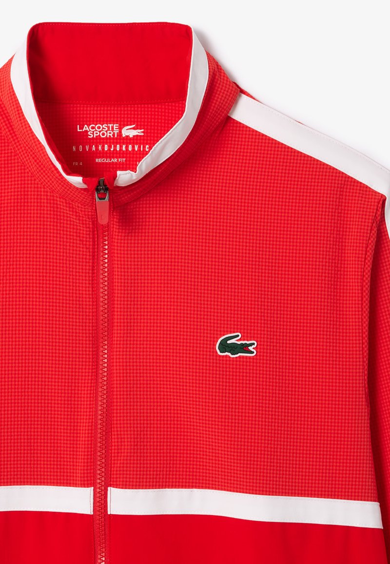 Veste zippée rouge avec un motif texturé, dotée d'accents blancs et d'un logo de crocodile vert sur la poitrine. Design à coupe régulière.