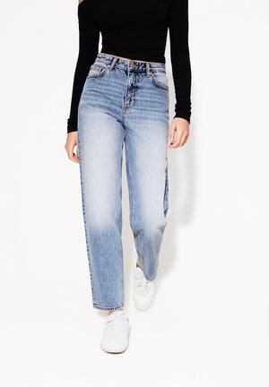 Osoba w jasnoniebieskich jeansach z wysokim stanem i prostymi nogawkami, czarnym topie z długim rękawem oraz białych trampkach, chodząca na białym tle.