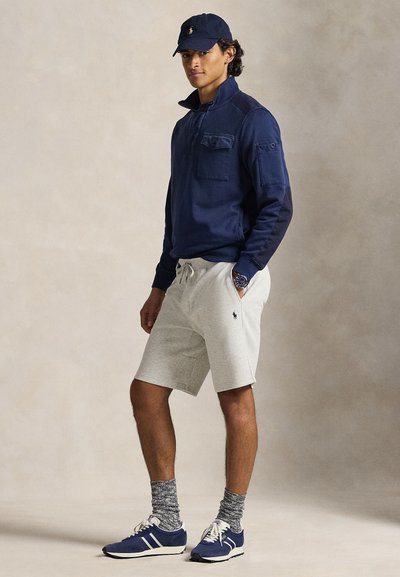 Ναυτικό μπλουζάκι με φερμουάρ, τσέπη στο στήθος, γκρίζες βερμούδες, μπλε αθλητικά παπούτσια με λευκές λεπτομέρειες και κάλτσες με σχέδια. Απλή, καθημερινή εμφάνιση.