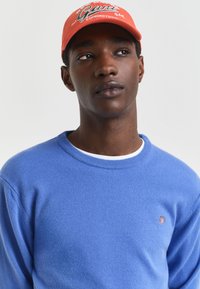 Junger Mann mit orangefarbener Baseballmütze und blauer Rundhals-Sweatshirt, der leicht zur Seite schaut und einen neutralen Ausdruck auf hellem Hintergrund hat.