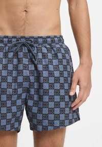 Calções de banho com padrão de quadriculado azul e preto e texto "Hugo". Apresentam cintura com cordão e tecido leve e texturizado.