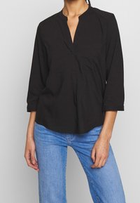 Camicia nera a maniche lunghe con colletto alto, chiusura a bottoni, due tasche sul petto e un fit rilassato, abbinata a jeans blu.