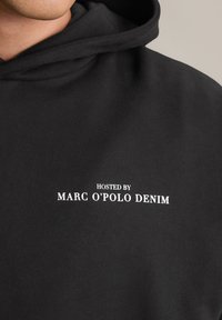 Zwarte hoodie gemaakt van zachte stof, met op de voorkant de witte tekst "GEPRESENTEERD DOOR MARC O'POLO DENIM".
