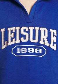 Blå pikétröja med krage, med "LEISURE" i krämfärgad brodyr och "1996" i vitt, centrerat på bröstet. Mjukt tyg med avslappnad passform.