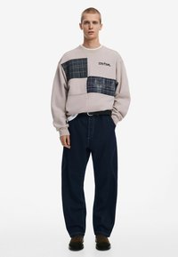 Sweatshirt in light beige with dark plaid patches, paired with loose dark denim pants. Features a relaxed fit and crew neckline.