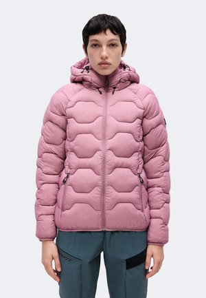 Jong persoon met kort donker haar, gekleed in een lichtroze doorgestikte gewatteerde jas met capuchon en donkergrijze broek tegen een witte achtergrond.