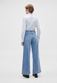 Hele sinise teksariidest laiad püksid kõrge vöökohaga, tagataskutega ja pruunist nahast plaastriga tagumisel vöökohal, kombineeritud heleda särgiga.