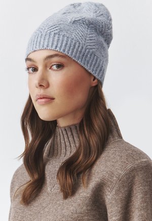 Lichtblauwe gebreide beanie met een gestructureerd patroon en ribbelband, gedragen bij een bruine trui met hoge kraag. Zacht en warm materiaal.