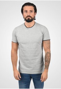Hombre con cabello oscuro y barba que lleva una camisa de manga corta gris claro y jeans azules, mostrando un tatuaje de un rostro barbudo en su antebrazo izquierdo.
