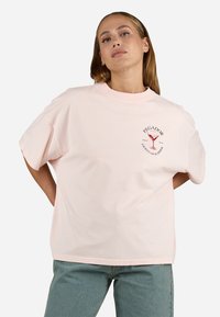 Lys rosa bomull t-skjorte med rund hals, korte ermer og en rød trykt logo med et cocktailglass på forsiden.