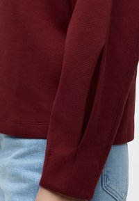Gros plan d’une personne portant un haut à manches longues bordeaux et un jean en denim bleu clair, montrant les détails de la manche et de la couture latérale.